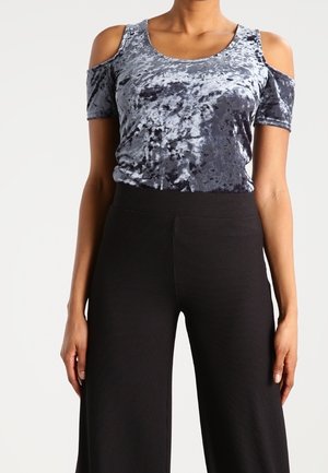 Vrouw draagt een blauw crushed velvet cold-shoulder top en zwarte wijde broek met hoge taille, staat met ontspannen armen langs haar zij.