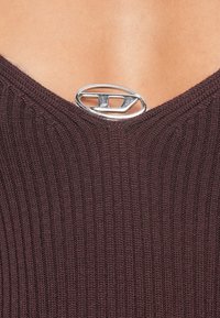 Tumši burgundijas krāsas adīts tops ar dziļu V veida izgriezumu un teksturētu ribojumu. Kakla izgriezumā ir sudraba ovāla piespraude ar logotipu.