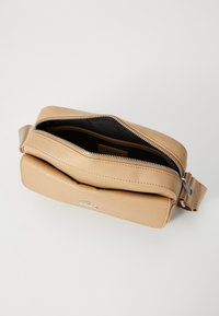 Beige teksturert skinnveggpose med frontlomme, sølvglidelås delvis åpen, og stoffrem, som viser svart innvendig fôr og Lacoste-merket.