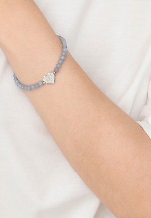 Wrist wearing a beaded gray bracelet with a silver geometric heart charm on a light skin tone against a white fabric background.