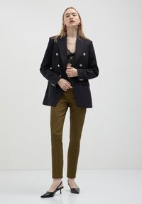Blazer blu navy doppiopetto con bottoni metallici, indossato sopra un top in pizzo nero. Pantaloni verdi oliva sartoriali e décolleté nere a punta.