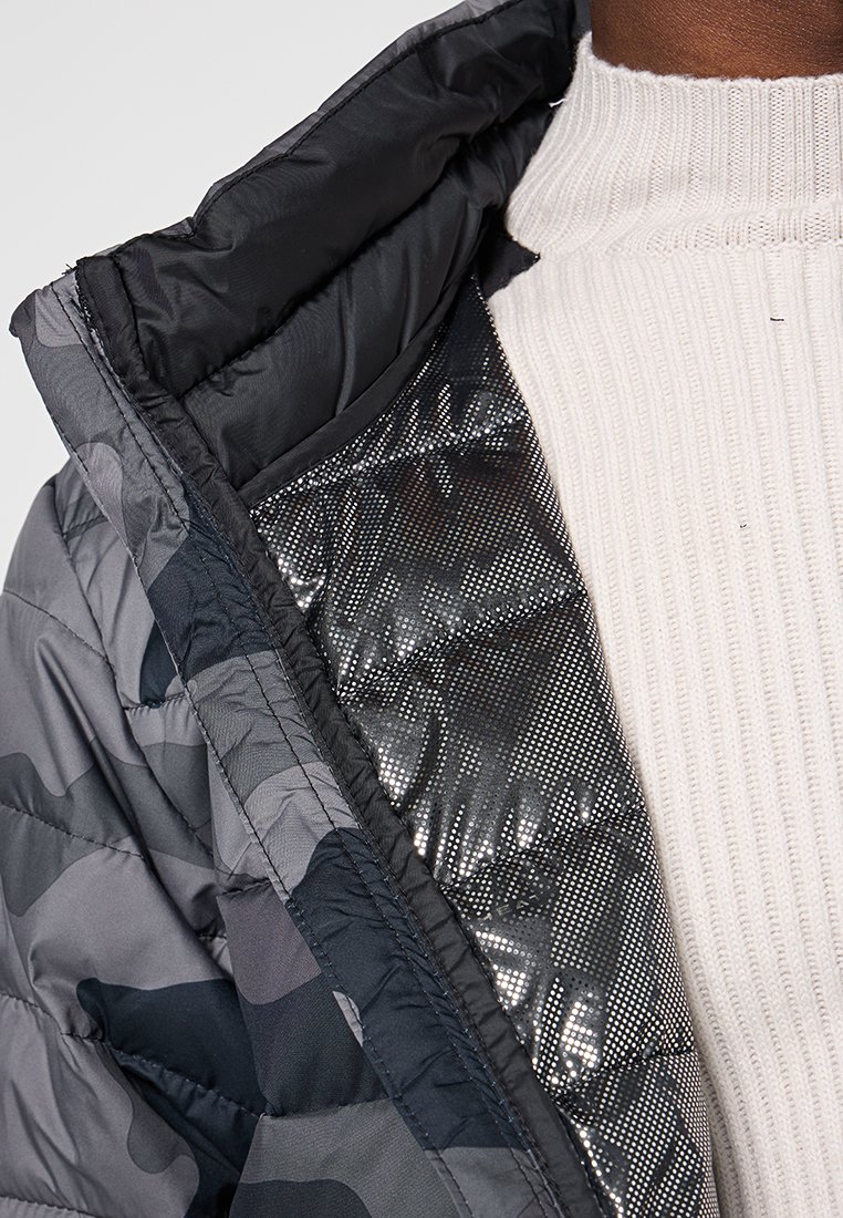 Chaqueta acolchada en patrón camuflaje gris, con un forro negro brillante. Suéter blanco acanalado visible debajo, con un cuello alto.