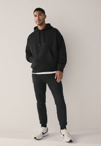 Sudadera con capucha negra con un bolsillo delantero, combinada con pantalones deportivos negros. El modelo lleva una camiseta blanca debajo y zapatillas claras con detalles.