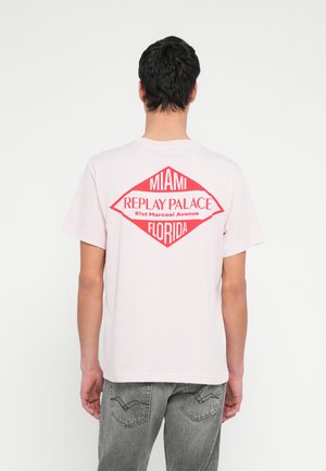 Man draagt een lichtroze T-shirt met een rode diamant en de tekst "Miami Replay Palace 81st Marcoai Avenue Florida" op de achterkant, grijze spijkerbroek.
