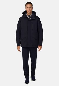 Mornarska izolirana jakna z visokim ovratnikom, nastavljivo kapuco, dvojnimi sprednjimi žepi in žepom na prsih z zadrgo; v kombinaciji z usklajenimi hlačami.