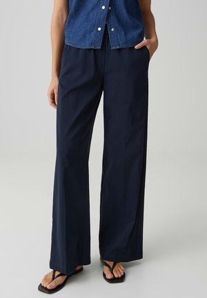 Person, der dunkelblaue Hosen mit weitem Bein, ein ärmelloses Jeanshemd mit Knöpfen und schwarze Flip-Flop-Sandalen trägt, steht mit einer Hand in der Hosentasche.