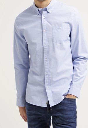 Uomo che indossa una camicia a maniche lunghe azzurro chiaro con bottoni e taschino sul petto, abbinata a jeans blu scuro, con una mano in tasca.