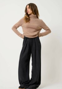 Beige Rollkragenpullover aus weichem Stoff, kombiniert mit hochgeschnittenen, weit geschnittenen schwarzen Hosen mit Falten und strukturiertem Material.