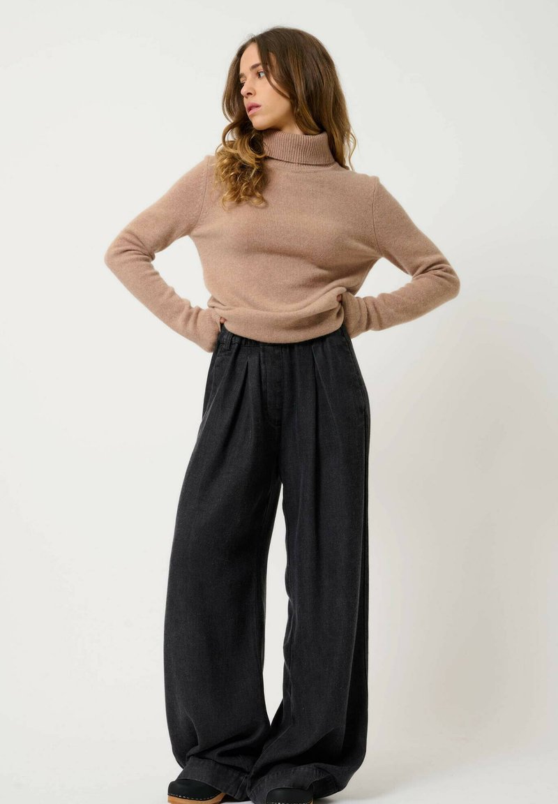Beige Rollkragenpullover aus weichem Stoff, kombiniert mit hochgeschnittenen, weit geschnittenen schwarzen Hosen mit Falten und strukturiertem Material.