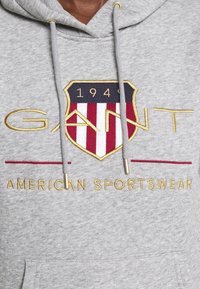 Sweat à capuche gris avec logo "GANT" brodé en or, écu rouge et blanc avec les textes "1949," et "AMERICAN SPORTSWEAR" en dessous.