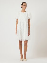 Άσπρο φόρεμα με υφασμάτινο λουλουδάτο μοτίβο, κοντά φουσκωτά μανίκια και εφαρμοστό κορμό που καταλήγει σε φουσκωτή φούστα. Κωνικά μπεζ παπούτσια.