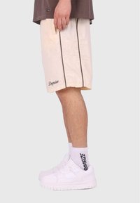 Helle beiges Shorts mit einer strukturierten Oberfläche, ausgestattet mit dunklen seitlichen Streifen und besticktem "Dropsize"-Branding, kombiniert mit weißen Sneakern und Socken.