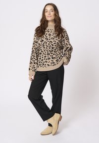 Beige tröja med svart leopardmönster, ribbat halslinning, avslappnad passform. Svarta byxor, beige ankelstövlar med en låg klack. Neutral bakgrund.