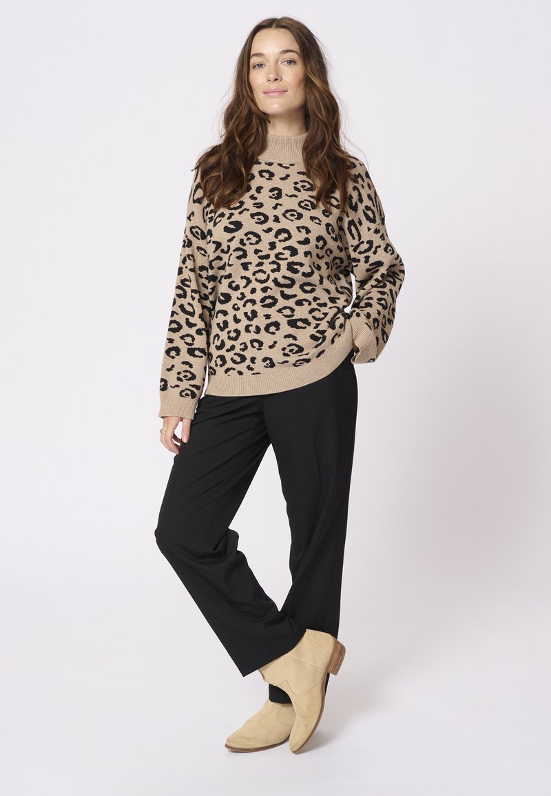 Beige tröja med svart leopardmönster, ribbat halslinning, avslappnad passform. Svarta byxor, beige ankelstövlar med en låg klack. Neutral bakgrund.