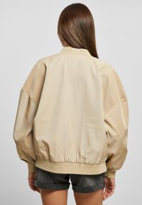 Giacca bomber beige con una finitura in satin liscio, maniche oversize, polsini e orlo a coste. Presenta un colletto classico e un fondo elasticizzato.