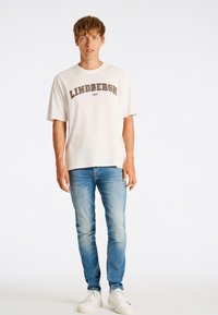 Camiseta de gran tamaño color crema con "LINDBERGH 1927" impreso en marrón oscuro. Combinada con jeans de color azul claro y zapatillas blancas, sobre un fondo simple.