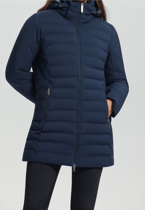 Femme portant une veste matelassée bleu marine mi-longue avec poches zippées sur les côtés, col montant et fermeture éclair devant, mains dans les poches.