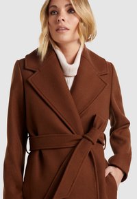 Brun uldfrakke med en indpakket design, brede revers og en belagt talje. Har en tekstureret cremefarvet turtleneck under.