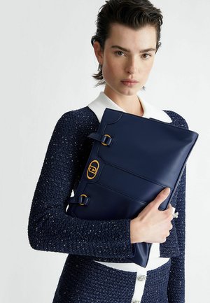 Clutch in pelle blu con dettagli in metallo dorato, caratterizzata da una forma strutturata e dettagli con fibbia, tenuta contro un capo in maglia blu a trama texture.