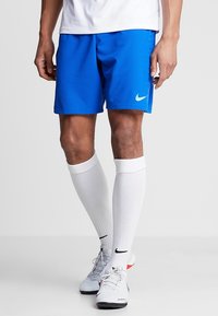 Pantalones cortos deportivos azules de Nike con un diseño texturizado, que presentan un logo verdoso en el lado izquierdo, combinados con calcetas blancas hasta la rodilla y zapatos grises.