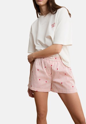 Frau trägt ein weißes, übergroßes T-Shirt mit rotem "Love is all"-Schriftzug und cremefarbene Shorts mit roten vertikalen Streifen und kleinen Herzmustern.