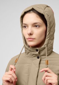 Beige gewatteerde jas met capuchon, voorzien van een ritssluiting en twee knopen. Inclusief verstelbare trekkoorden met houten accenten.