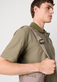 Leichte graue Schultertasche mit "BOSS"-Logo, ausgestattet mit einem robusten Tragegurt und kontrastierendem Textilgewebe. Getragen von einem Modell in einem grünen Poloshirt.