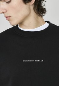 Sweatshirt noir en coton avec un col rond, portant le texte blanc "Denmark Street - London UK" centré sur la poitrine.