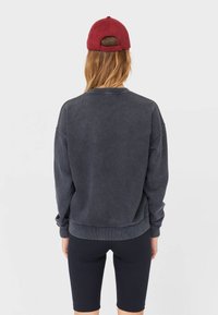 Donkergrijze sweatshirt met een ontspannen ontwerp, geribbelde mouw- en onderrand. Gecombineerd met zwarte fietsbroek en een bordeauxrode honkbalpet.
