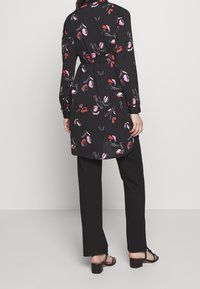 Svart skjortklänning med blommönster i rosa och rött. Långa ärmar, knäppning fram, knytning i midjan och sidoklyftor. Bärs med svarta byxor och sandaler.