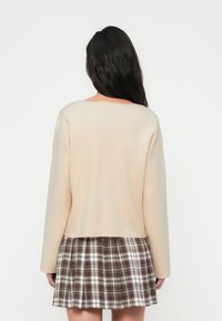 Μπεζ μακρυμάνικο τοπ από απαλό ύφασμα, συνδυασμένο με καρό φούστα σε καφέ και λευκούς τόνους, δημιουργώντας ένα στρωματοποιημένο look.