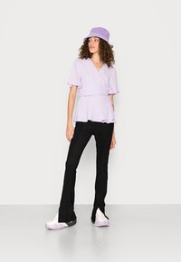 Mulher vestindo uma blusa envolvente lilás clara, um chapéu de balde combinando, calças flare pretas e tênis brancos com solas roxas, contra um fundo branco.