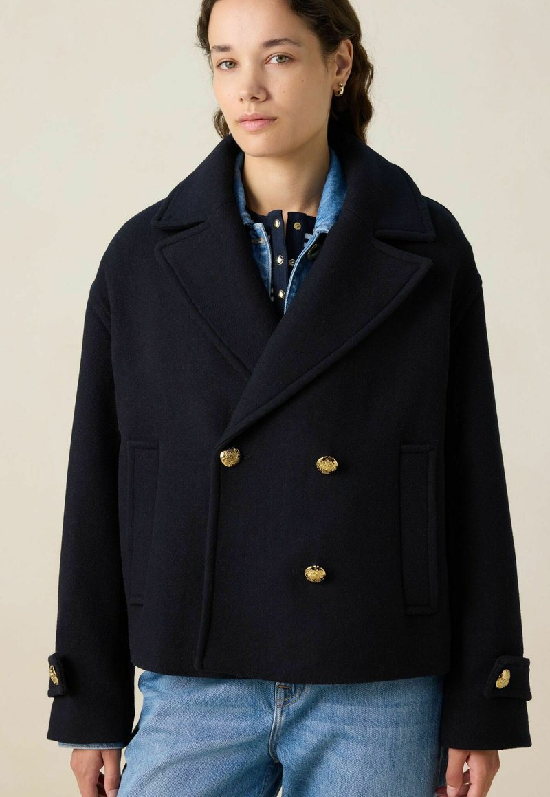 Manteau en laine bleu marine à double boutonnage avec un large col, des boutons dorés et des poches latérales ; porté sur une chemise en denim bleue à pois.