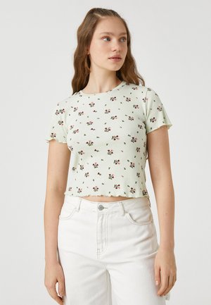 Jasnozielony, prążkowany crop top z kwiatowym wzorem w czerwonych i zielonych akcentach. Posiada okrągły dekolt i krótkie rękawy z falbaniastymi krawędziami.