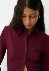 Burgundy skjorta med knapp-up och krage, med en figursydd design och jämn textur. Sju knappar synliga fram.