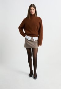 Brązowy wełniany sweter z golfem i prążkowanymi mankietami, nałożony na białą koszulę z kołnierzykiem, w zestawieniu z beżową mini spódnicą i czarnymi szpilkami.