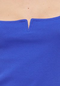 Κοντινό πλάνο ενός μπλε ριμπ τοπ με έξω από τους ώμους και μικρό κεντρικό άνοιγμα στη λαιμόκοψη, που αφήνει να φανεί το ανώτερο μέρος του στήθους.