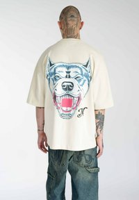 Creme oversized t-shirt med et blåt og rødt ulvegrafik på ryggen, lavet af bomuld, med korte ærmer og en afslappet pasform.