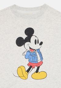 Topolino che indossa una giacca di denim blu, pantaloni rossi e grandi scarpe gialle, che cammina con le mani dietro la schiena su uno sfondo di tessuto grigio chiaro.