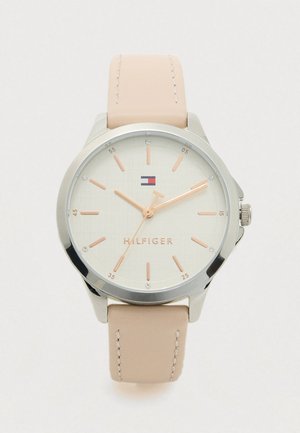 Okrogla srebrna ura na zapestju s bež usnjeno naramnico, svetlim številčnikom, rožnatimi zlatimi kazalci in oznakami ur ter majhnim rdečim, belim in modrim logotipom pod številko 12.