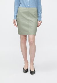 Saia verde de couro sintético com design ajustado, comprimento até ao joelho e textura suave. Combinada com um cardigan azul-claro e sapatilhas pretas.