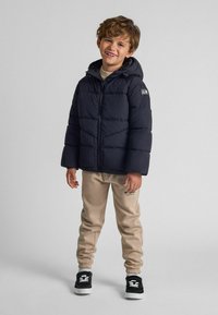 Tumši zils pūkains jaka ar kapuci un rāvējslēdzēju, bēšas treniņbikses un melnas sporta apavus. Iekļauj logotipu uz piedurknes un elastīgus neplīstošus rokas .