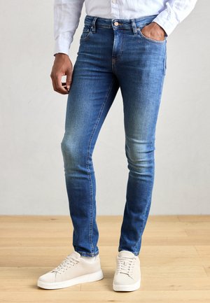 Jeans di denim blu aderenti con una tonalità sfumata e design classico a cinque tasche. Indossati con sneakers bianche e una camicia di colore chiaro.