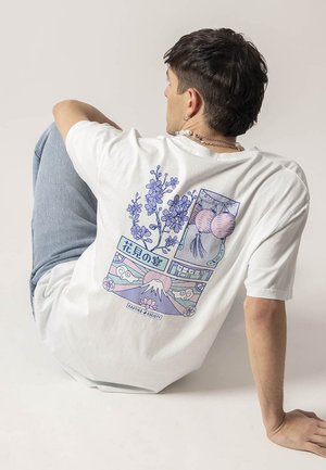 Λευκό βαμβακερό t-shirt με ζωντανό μπλε γραφικό σχέδιο στην πλάτη, που απεικονίζει λουλούδια και παραδοσιακά μοτίβα, με χαλαρή εφαρμογή.