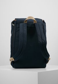 Granatowy plecak z materiału o prostokątnym kształcie, z beżowymi paskami i akcentami w postaci zamków błyskawicznych. Posiada wyściełane ramiączka oraz uchwyt na górze do noszenia.