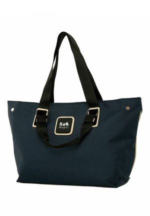Sac fourre-tout en toile bleu marine avec des poignées noires, présentant une étiquette carrée avec la marque et des accents de quincaillerie argentée. Design grand et spacieux.