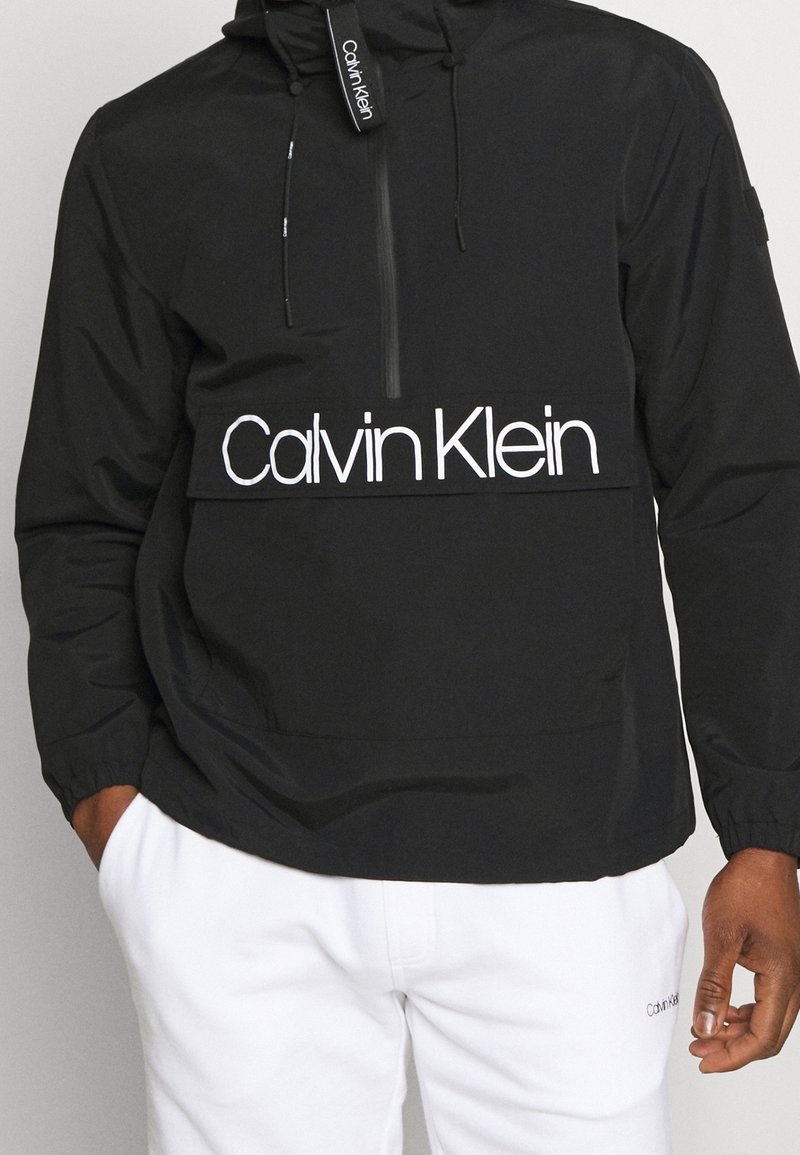 Personne portant un sweat à capuche noir Calvin Klein avec fermeture éclair frontale et pantalon blanc Calvin Klein, mains partiellement visibles, debout sur un fond neutre.