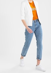 Witte getailleerde blazer over een oranje T-shirt met grafische print, gecombineerd met lichtblauwe high-waisted jeans en witte sneakers.