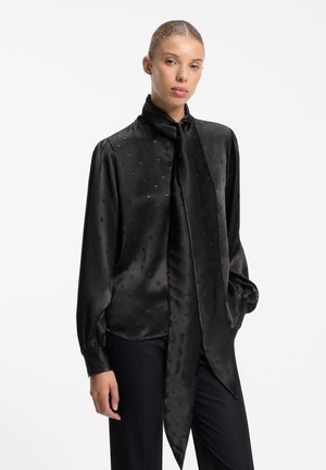 Woman wearing a black satin blouse with subtle polka dots and a long neck tie, paired with black pants against a plain white background.