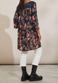 Marinblommig tunika med långa ärmar och veckad bakdesign, kombinerad med vita figurvänliga byxor och svarta knähöga stövlar.